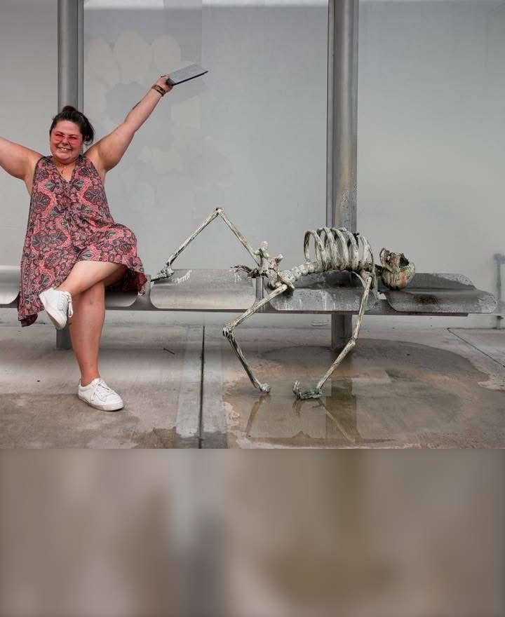 Crystal Adair-Benning, arms in the air on a bench beside a skeleton that has been waiting a very long time