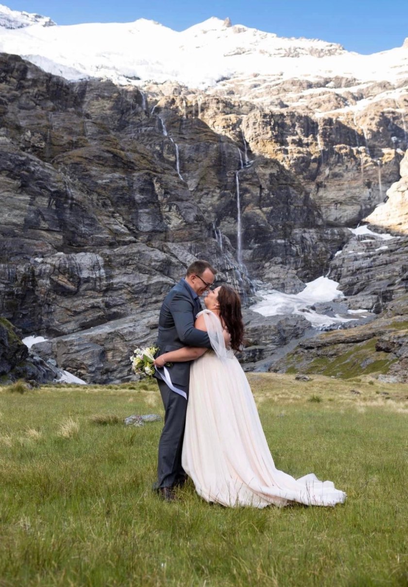 Crystal Adair-Benning and her husband kissing in a meadow beneath a glacier and waterfalls