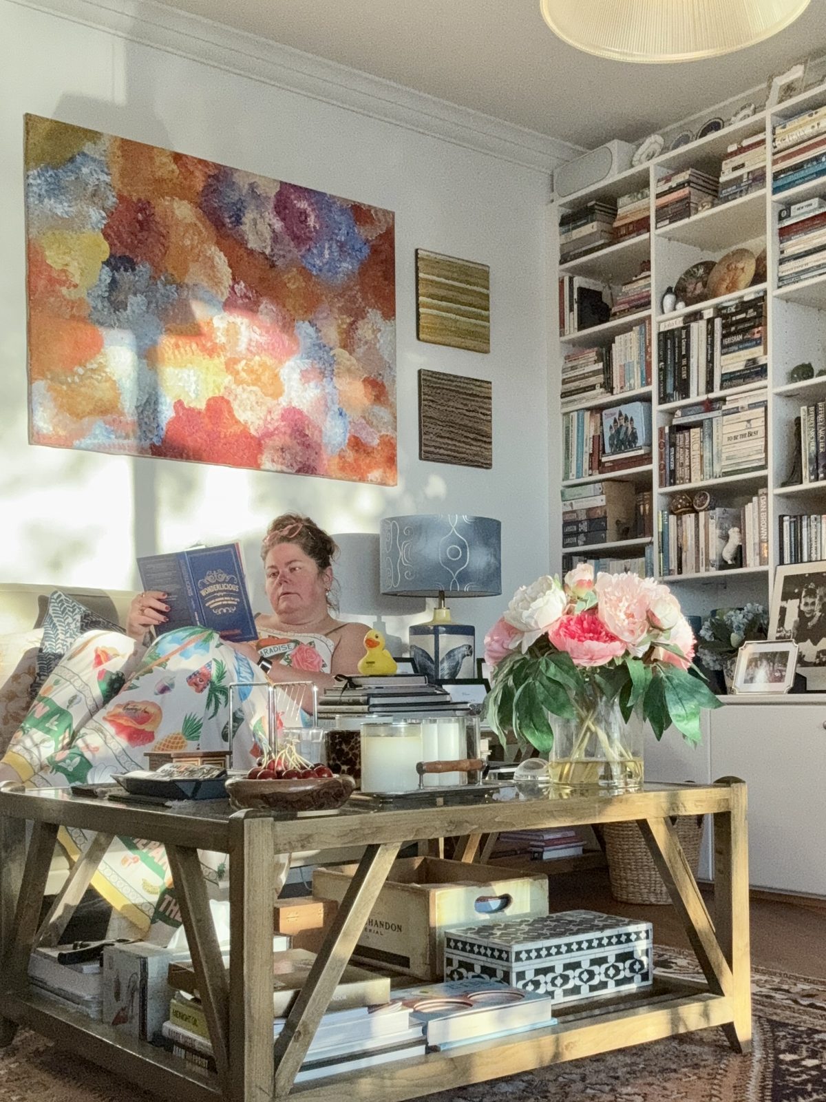 Crystal Adair-Benning reading on a sofa beside peonies and bookshelves in Sydney